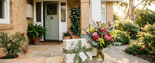 A beautifully wrapped gift box with a green ribbon sitting on a suburban Australian doorstep next to a bouquet of native flowers like Banksias and Proteas in the morning sun.