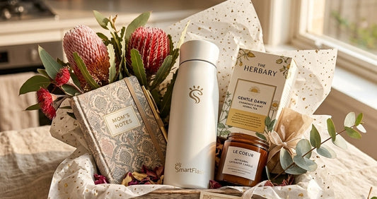 A luxurious DIY Mother's Day hamper featuring a Pearl White SmartFlask water bottle, herbal teas, a scented candle, and a journal nestled with native Australian flowers in a wooden tray.