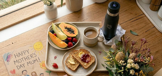 A black SmartFlask UV water bottle on a sunlit wooden kitchen bench next to a Mother's Day breakfast tray with fruit and toast.
