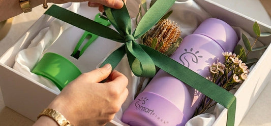 Hand tying a green ribbon on a luxury gift box containing a purple SmartFlask, native flowers, and green accessories, under warm morning light.