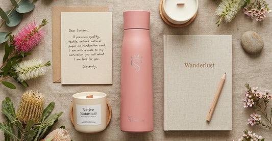 Flat lay of under $50 gifts, including a SmartFlask water bottle, card, candle, native flowers, and a journal on a linen surface.